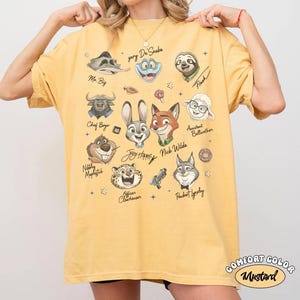 May include: Mustard-colored Comfort Colors t-shirt featuring cartoon character faces from Zootopia, including Judy Hopps and Nick Wilde. The shirt has the text "Joy Happy" and character names. The shirt is a relaxed fit.