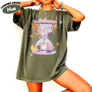 May include: Moss green t-shirt featuring a whimsical hourglass design with cartoon characters, flowers, and stars. The design includes a moon, hearts, and a variety of cute creatures. The shirt has the words "Comfort Colors" above the word "Moss".