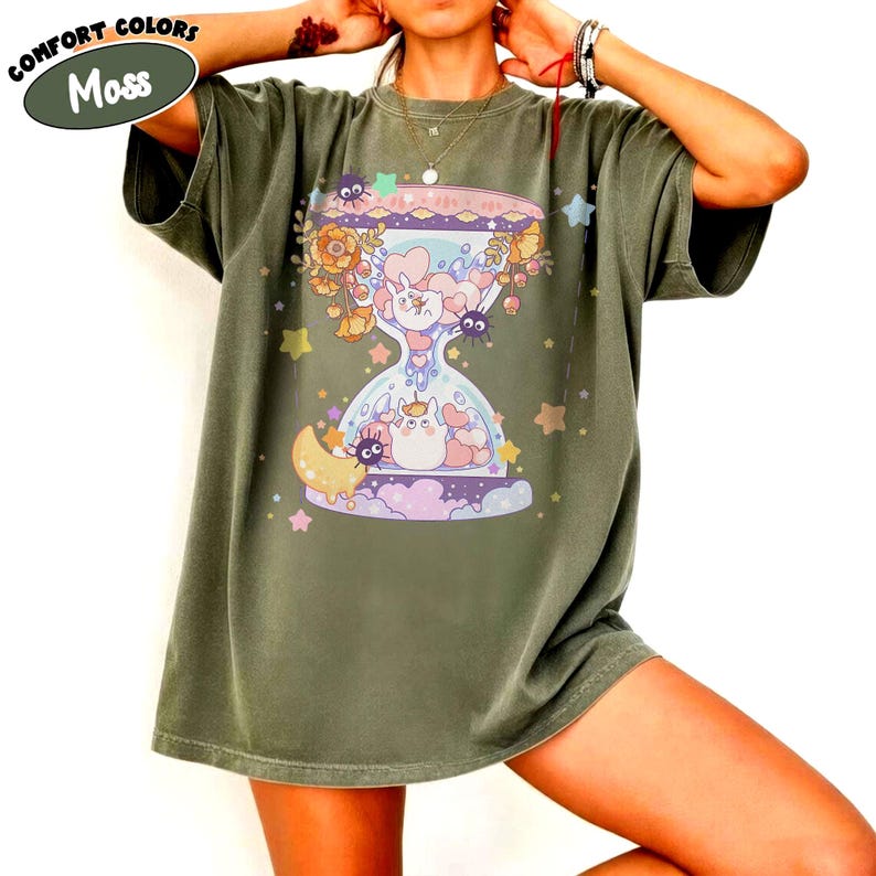 May include: Moss green t-shirt featuring a whimsical hourglass design with cartoon characters, flowers, and stars. The design includes a moon, hearts, and a variety of cute creatures. The shirt has the words "Comfort Colors" above the word "Moss".