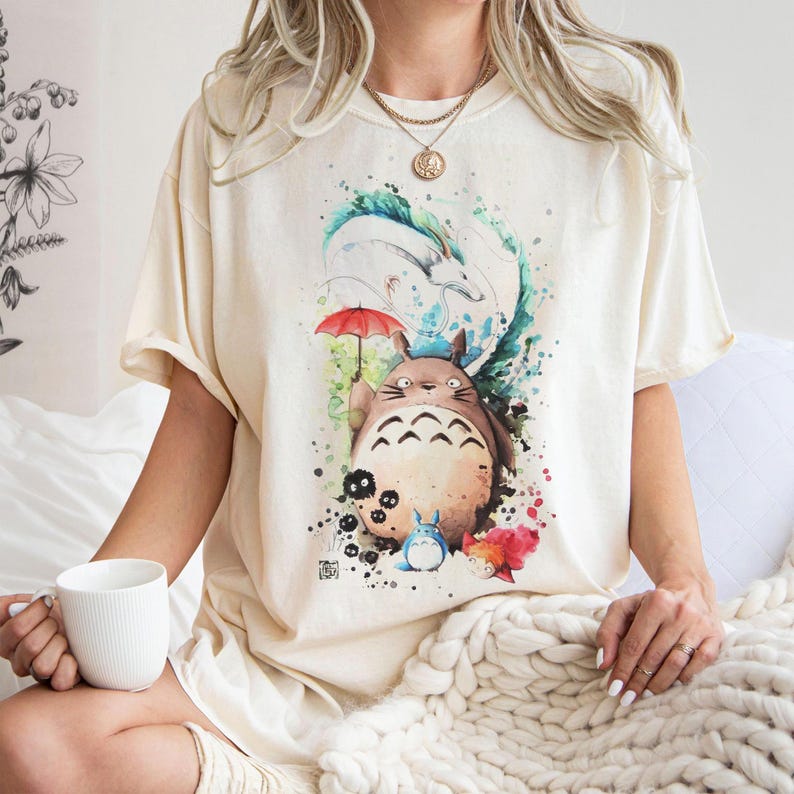 以下が含まれることがあります： 人気アニメのキャラクターの水彩イラストが描かれたクリーム色のTシャツ。赤い傘を持った大きくて茶色い毛むくじゃらの生き物、他のキャラクター、カラフルなアクセントがデザインに含まれています。