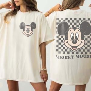May include: Ivory-colored t-shirt featuring a distressed Mickey Mouse graphic on the front and back. The back design includes a checkered pattern and the words "Mickey Mouse". The shirt has short sleeves and a relaxed fit.