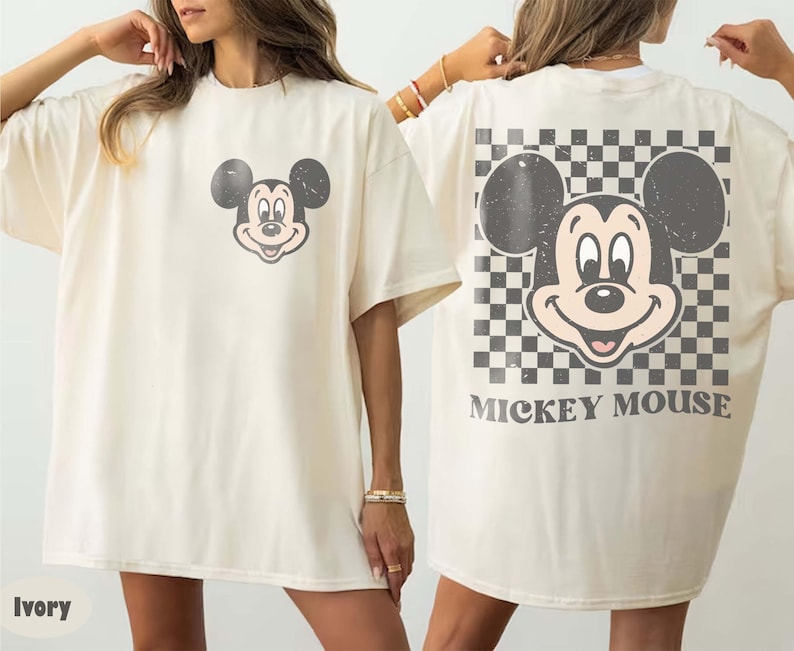 May include: Ivory-colored t-shirt featuring a distressed Mickey Mouse graphic on the front and back. The back design includes a checkered pattern and the words "Mickey Mouse". The shirt has short sleeves and a relaxed fit.