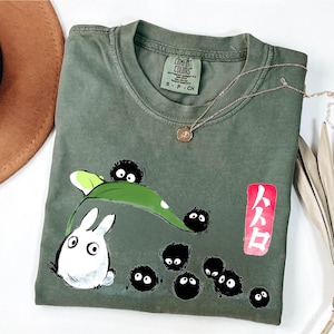 May include: Olive green Comfort Colors t-shirt featuring a cartoon design with a white rabbit-like creature, black soot sprites, and a green leaf. A gold necklace with a pendant is draped across the shirt. The shirt also has Japanese text on a red rectangle.