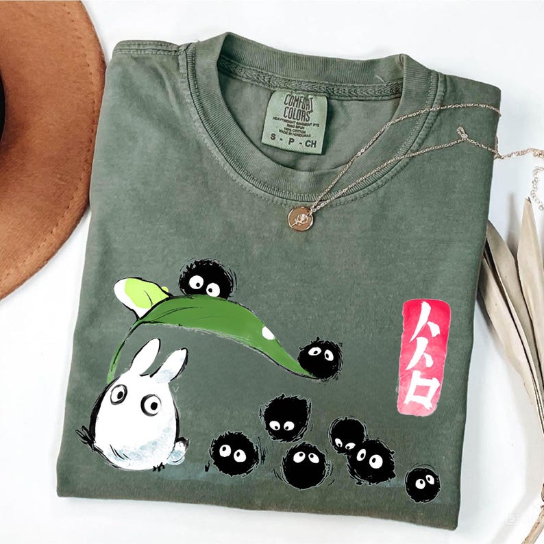 May include: Olive green Comfort Colors t-shirt featuring a cartoon design with a white rabbit-like creature, black soot sprites, and a green leaf. A gold necklace with a pendant is draped across the shirt. The shirt also has Japanese text on a red rectangle.
