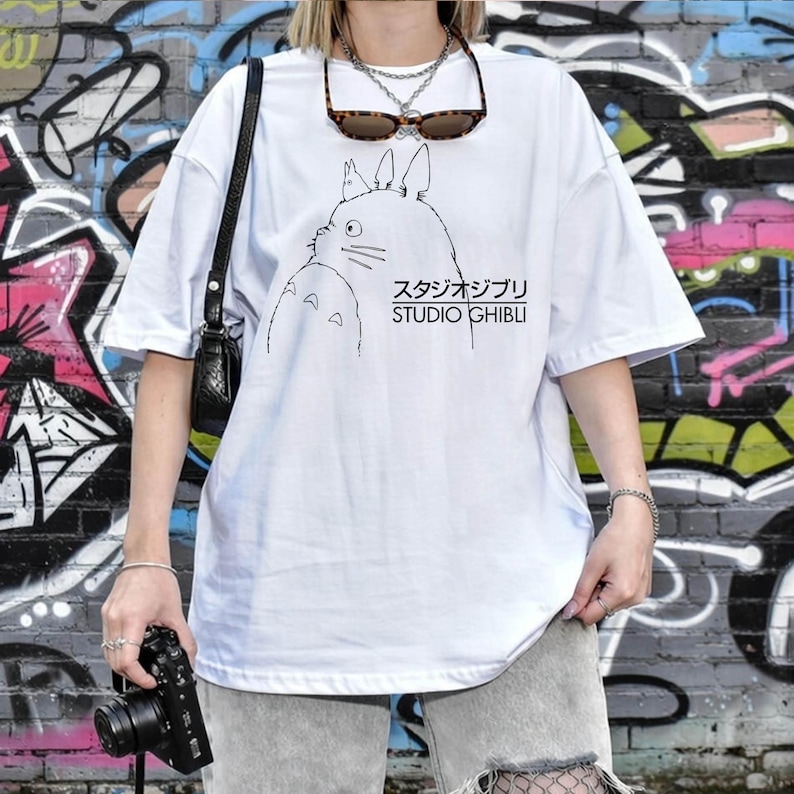 May include: White t-shirt featuring a black line drawing of a character from Studio Ghibli, with Japanese text and "STUDIO GHIBLI" printed below. The shirt has short sleeves and a relaxed fit.