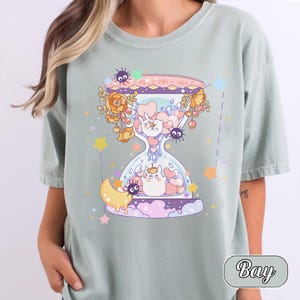May include: Sage green t-shirt featuring a whimsical hourglass design. The hourglass is filled with cartoon characters, hearts, and flowers. The design includes a crescent moon, stars, and the word "Bay" in a rounded font.