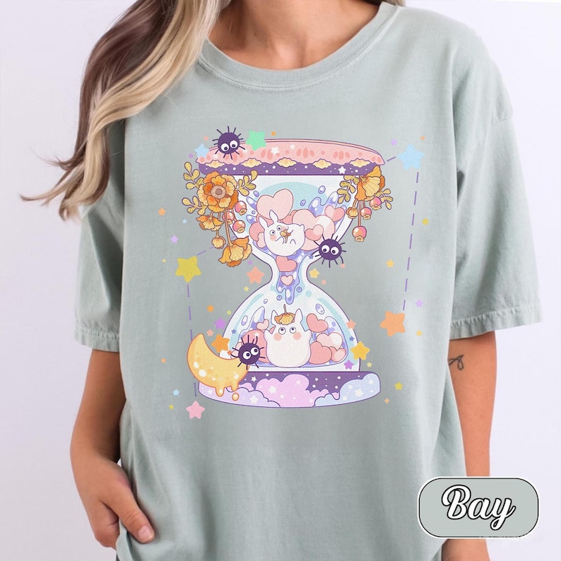 May include: Sage green t-shirt featuring a whimsical hourglass design. The hourglass is filled with cartoon characters, hearts, and flowers. The design includes a crescent moon, stars, and the word "Bay" in a rounded font.
