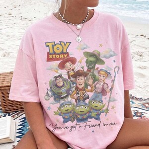 May include: A light pink short-sleeve t-shirt featuring a colorful graphic print of Toy Story characters including Woody, Buzz Lightyear, Jessie, Rex, Bo Peep, and the Aliens. The text reads Toy Story and You've got a Friend in me.