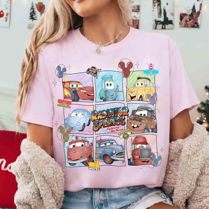 Puede incluir: Camiseta rosa claro con un dise&ntilde;o gr&aacute;fico colorido de personajes de la pel&iacute;cula Cars. El dise&ntilde;o incluye im&aacute;genes de varios coches y el texto "Radiator Springs". La camiseta tiene cuello redondo y mangas cortas.