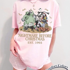 May include: A light pink short-sleeve t-shirt featuring a graphic print of characters from The Nightmare Before Christmas, including Jack Skellington, Sally, and Oogie Boogie, with the text NIGHTMARE BEFORE CHRISTMAS EST. 1993 printed in gold.