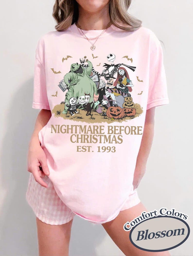 May include: A light pink short-sleeve t-shirt featuring a graphic print of characters from The Nightmare Before Christmas, including Jack Skellington, Sally, and Oogie Boogie, with the text NIGHTMARE BEFORE CHRISTMAS EST. 1993 printed in gold.