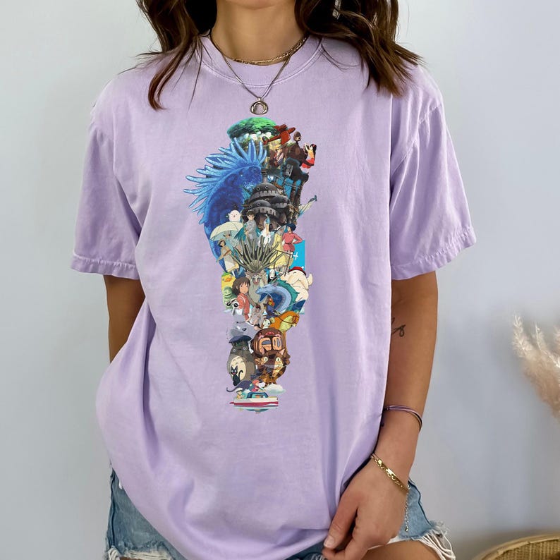 May include: A light purple t-shirt featuring a colorful collage of anime characters and scenes. The graphic is vertically oriented and includes images of various animated figures and landscapes. The shirt has short sleeves and a relaxed fit.