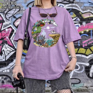 May include: A lilac-colored t-shirt with a colorful circular graphic featuring animated characters and landscapes. The text "ORCHID COMFORT COLORS" is printed at the top left. The shirt is worn with gray shorts and a black purse.