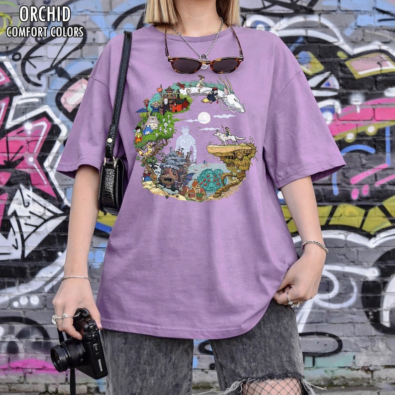 May include: A lilac-colored t-shirt with a colorful circular graphic featuring animated characters and landscapes. The text "ORCHID COMFORT COLORS" is printed at the top left. The shirt is worn with gray shorts and a black purse.