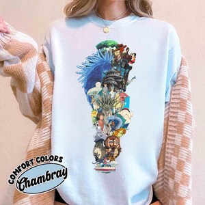 May include: Light blue sweatshirt featuring a colorful graphic design of various anime characters. The design includes a blue dragon, forest, and several figures. The sweatshirt has the text "Comfort Colors Chambray" in a circular design.
