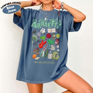 May include: Blue jean comfort colors t-shirt with a graphic from "The Secret World of Arrietty". The design features various illustrations including insects, food, and tools. The text is in English and Japanese.
