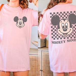 May include: A light pink t-shirt featuring a distressed Mickey Mouse graphic on the front and back. The back design includes a checkered pattern and the words "Mickey Mouse". The shirt is a casual, oversized style.