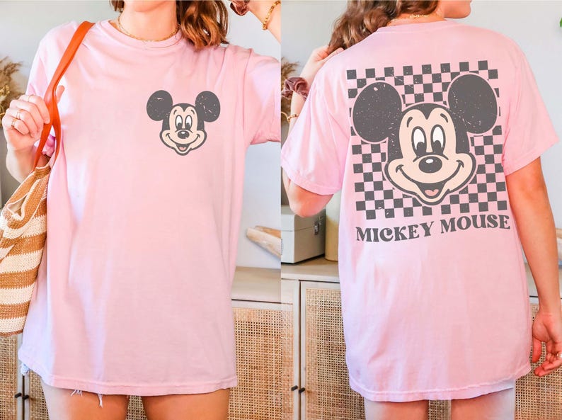May include: A light pink t-shirt featuring a distressed Mickey Mouse graphic on the front and back. The back design includes a checkered pattern and the words "Mickey Mouse". The shirt is a casual, oversized style.