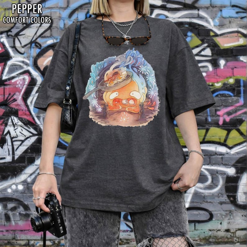 Puede incluir: Camiseta gris oscuro con un gr&aacute;fico de dibujos animados colorido de un personaje cocinando en una sart&eacute;n. La camiseta tiene el texto "PEPPER COMFORT COLORS" en la parte superior. La camiseta se usa frente a una pared de graffiti.