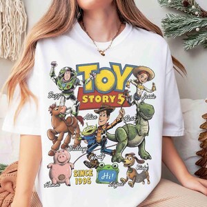 May include: A white t-shirt featuring a colorful graphic print of Toy Story characters including Buzz Lightyear, Woody, Jessie, Bullseye, Rex, Hamm, Slinky, and an Alien. The design includes the text Toy Story 5, Since 1995, and character names.