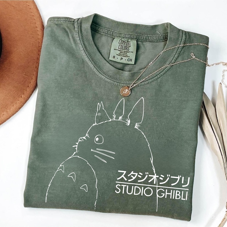May include: An olive green Comfort Colors t-shirt featuring a white graphic of a cartoon character and the words "STUDIO GHIBLI" in English and Japanese. A gold necklace with a pendant rests on the shirt.