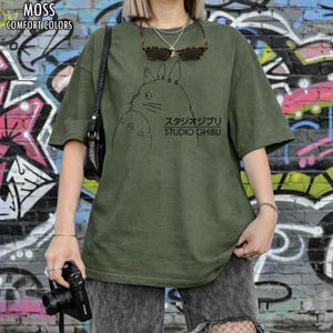 May include: Moss green t-shirt with a black graphic of a cartoon character and the text "スタジオジブリ STUDIO GHIBLI". The shirt has short sleeves and a relaxed fit. The words "MOSS" and "COMFORT COLORS" are printed at the top.