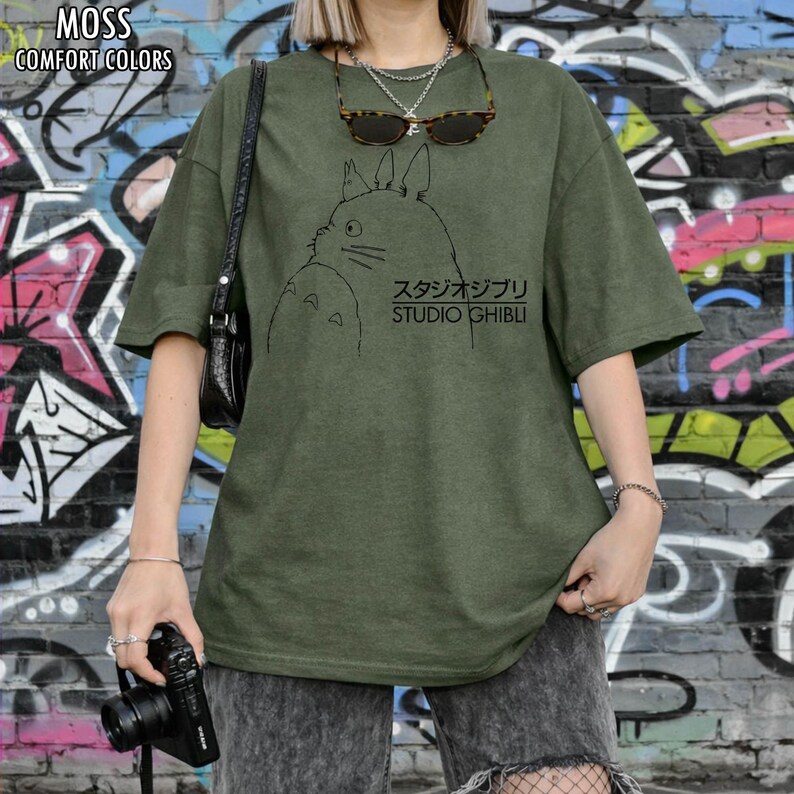 May include: Moss green t-shirt with a black graphic of a cartoon character and the text "スタジオジブリ STUDIO GHIBLI". The shirt has short sleeves and a relaxed fit. The words "MOSS" and "COMFORT COLORS" are printed at the top.