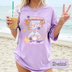 May include: A lilac-colored t-shirt featuring a whimsical hourglass design with cartoon characters, flowers, and stars. The text "Orchid" is in a cursive font at the bottom right. The shirt has short sleeves and a relaxed fit.