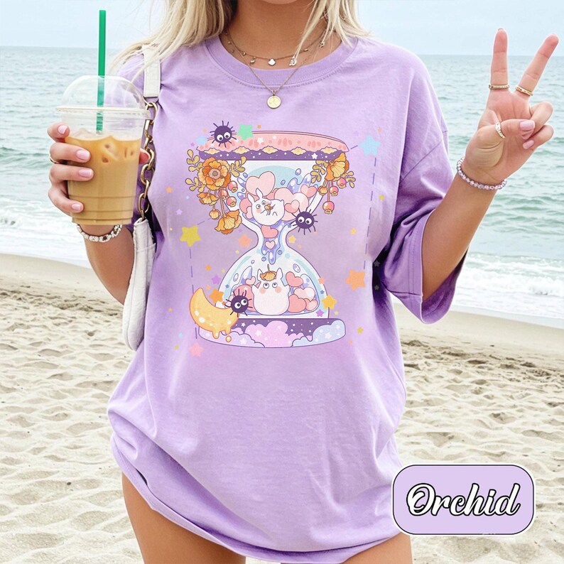 May include: A lilac-colored t-shirt featuring a whimsical hourglass design with cartoon characters, flowers, and stars. The text "Orchid" is in a cursive font at the bottom right. The shirt has short sleeves and a relaxed fit.