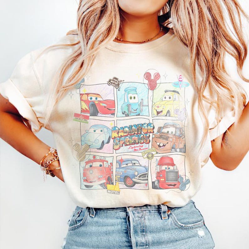 Puede incluir: Camiseta color crema con un gr&aacute;fico colorido de personajes de la pel&iacute;cula Cars. El dise&ntilde;o incluye varios coches en un estilo de c&oacute;mic, con las palabras "Radiator Springs" y "Lightning McQueen".