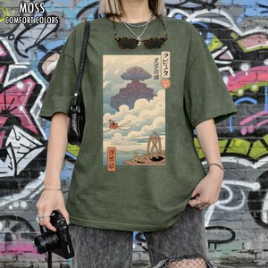 May include: Moss green t-shirt with a Japanese anime-style graphic. The graphic features a floating castle, characters, and Japanese text. The shirt has the words "MOSS COMFORT COLORS" printed at the top.