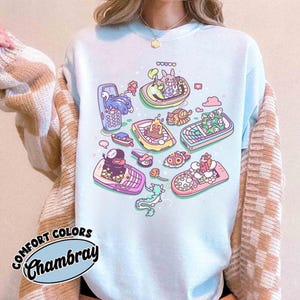 K&ouml;nnte beinhalten: Hellblaues Comfort Colors Chambray T-Shirt mit einem skurrilen Design von Zeichentrickfiguren in Booten in Form von Retro-Handys. Das Design umfasst verschiedene Farben und Details.