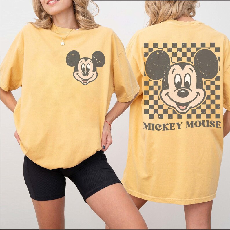 May include: Mustard yellow t-shirt featuring a distressed Mickey Mouse graphic on the front and back. The back design includes a checkered pattern and the words "Mickey Mouse". The shirt has a relaxed fit and short sleeves.