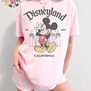May include: A light pink t-shirt featuring a graphic of Mickey Mouse giving a peace sign in front of a castle silhouette. The word "Disneyland" is arched above the character, with "California" below. The shirt also has the text "Est 1955".