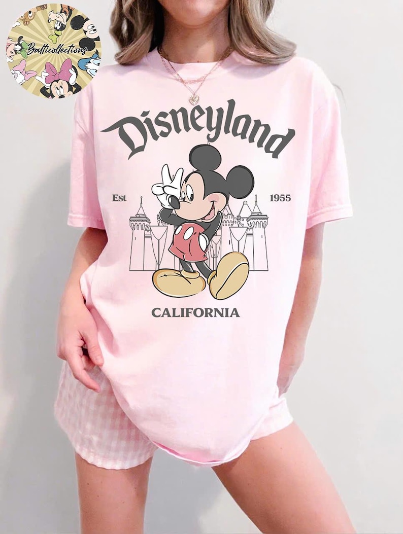 May include: A light pink t-shirt featuring a graphic of Mickey Mouse giving a peace sign in front of a castle silhouette. The word "Disneyland" is arched above the character, with "California" below. The shirt also has the text "Est 1955".