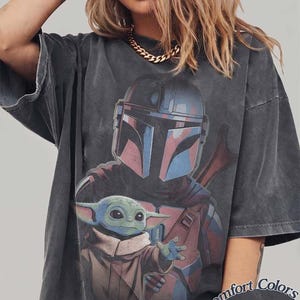 May include: A charcoal gray oversized t-shirt featuring a graphic print of a helmeted warrior and a small green creature with large ears. The shirt has a faded, washed-out appearance and includes the text Comfort Colors Pepper in the bottom corner.