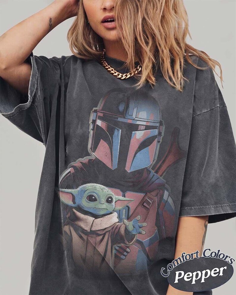 May include: A charcoal gray oversized t-shirt featuring a graphic print of a helmeted warrior and a small green creature with large ears. The shirt has a faded, washed-out appearance and includes the text Comfort Colors Pepper in the bottom corner.