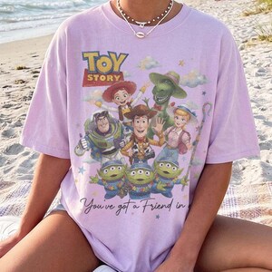 May include: A light purple short-sleeve t-shirt featuring a graphic print of Toy Story characters including Woody, Buzz Lightyear, Jessie, Rex, Bo Peep, and the Aliens. The text reads Toy Story and You've got a Friend in Me with small stars.