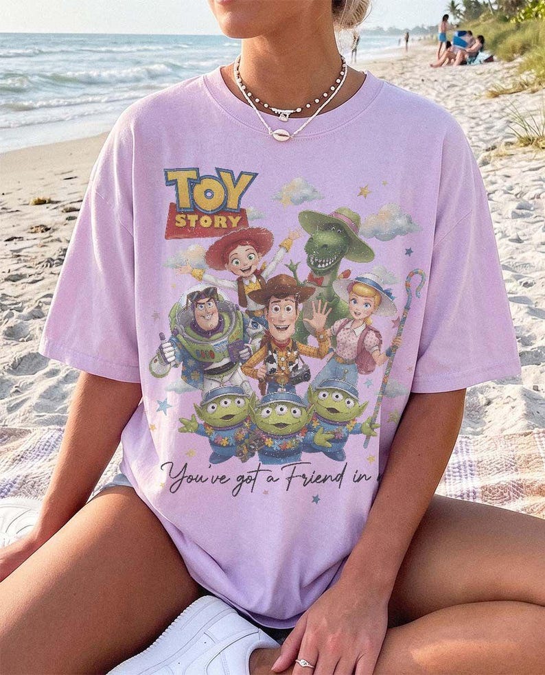 May include: A light purple short-sleeve t-shirt featuring a graphic print of Toy Story characters including Woody, Buzz Lightyear, Jessie, Rex, Bo Peep, and the Aliens. The text reads Toy Story and You've got a Friend in Me with small stars.