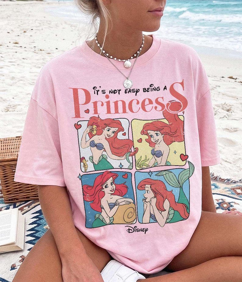 May include: A light pink short-sleeve t-shirt featuring a graphic print of the character Ariel from The Little Mermaid in four separate panels. The text "it's not easy being a Princess" is printed above the panels, with the Disney logo below.