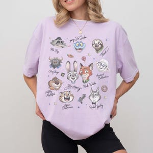 May include: A light purple t-shirt featuring cartoon characters from a movie. The shirt has illustrations of a bunny, fox, sloth, and other characters. The names of the characters are written below them.