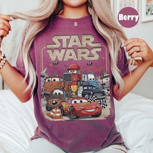 Puede incluir: Camiseta color baya con un dise&ntilde;o gr&aacute;fico que muestra las palabras "STAR WARS" sobre una imagen de dibujos animados de veh&iacute;culos de la pel&iacute;cula "Cars". La imagen incluye a Rayo McQueen, Mate y otros personajes, con una bandera a cuadros y un trofeo.