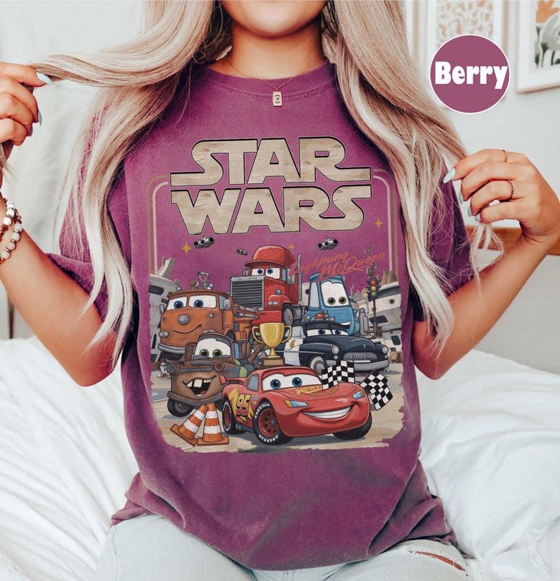 Puede incluir: Camiseta color baya con un dise&ntilde;o gr&aacute;fico que muestra las palabras "STAR WARS" sobre una imagen de dibujos animados de veh&iacute;culos de la pel&iacute;cula "Cars". La imagen incluye a Rayo McQueen, Mate y otros personajes, con una bandera a cuadros y un trofeo.