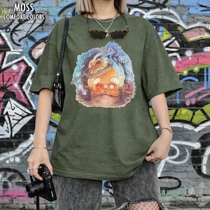 Puede incluir: Camiseta verde oliva con un dibujo animado de un personaje cocinando en una sart&eacute;n. La camiseta tiene las palabras "MOSS COMFORT COLORS" impresas en la parte superior. La modelo lleva gafas de sol y un collar plateado.
