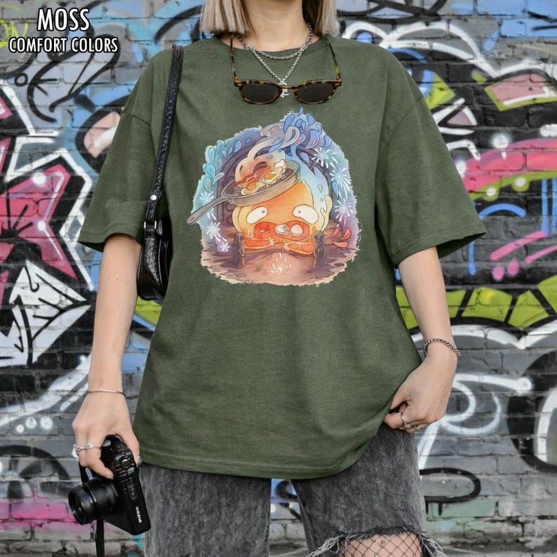 Puede incluir: Camiseta verde oliva con un dibujo animado de un personaje cocinando en una sart&eacute;n. La camiseta tiene las palabras "MOSS COMFORT COLORS" impresas en la parte superior. La modelo lleva gafas de sol y un collar plateado.