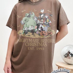 May include: A brown short-sleeve t-shirt featuring a graphic print of characters from The Nightmare Before Christmas, including Jack Skellington, Sally, and Oogie Boogie. The text reads NIGHTMARE BEFORE CHRISTMAS EST. 1993. A Comfort Colors Espresso label is visible.