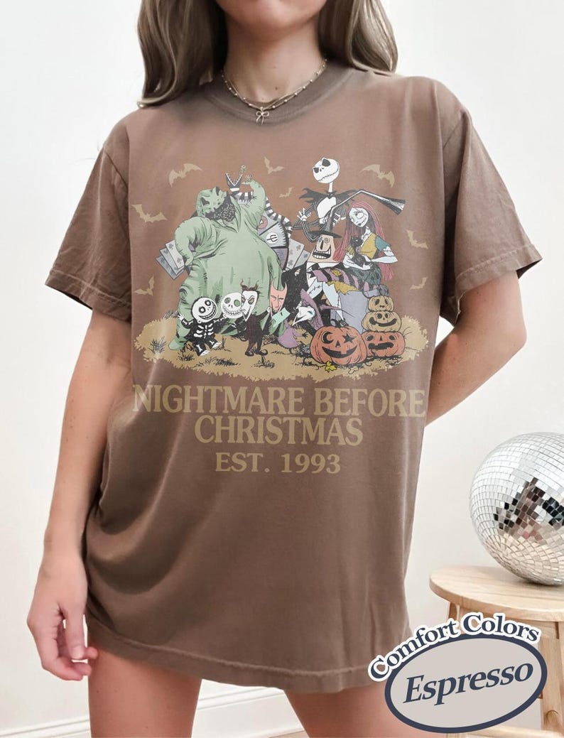 May include: A brown short-sleeve t-shirt featuring a graphic print of characters from The Nightmare Before Christmas, including Jack Skellington, Sally, and Oogie Boogie. The text reads NIGHTMARE BEFORE CHRISTMAS EST. 1993. A Comfort Colors Espresso label is visible.