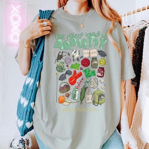May include: A light green t-shirt with the text "THE SECRET WORLD OF ARRIETY" in green, along with various illustrated elements. The illustrations include insects, berries, and jars. The shirt is being worn by a person.