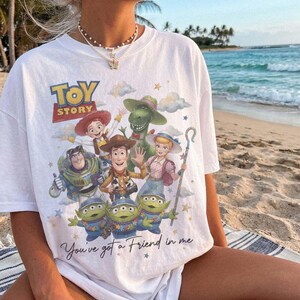 May include: A white t-shirt featuring a colorful graphic print of Toy Story characters including Woody, Buzz Lightyear, Jessie, Bo Peep, Rex, and three aliens. The text Toy Story and You've got a Friend in me are printed on the front.