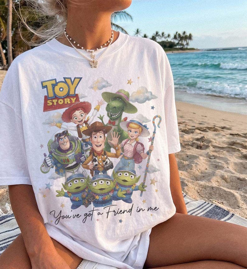 May include: A white t-shirt featuring a colorful graphic print of Toy Story characters including Woody, Buzz Lightyear, Jessie, Bo Peep, Rex, and three aliens. The text Toy Story and You've got a Friend in me are printed on the front.